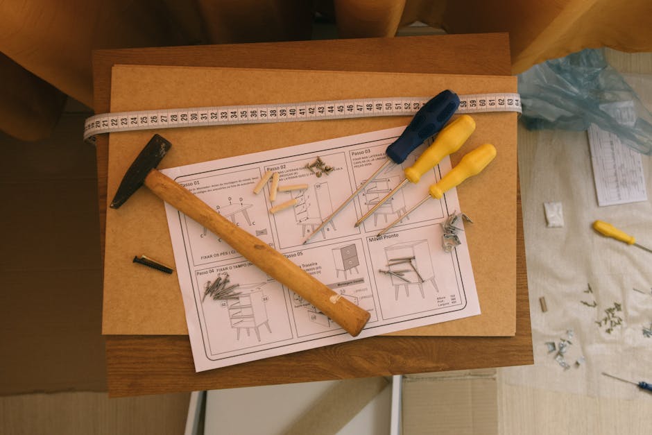 A man with dark hair and a beard, wearing a white t-shirt, is using a screwdriver to dismantle a white baby cot or crib in a well-lit indoor setting. He appears focused as he works on removing a screw
