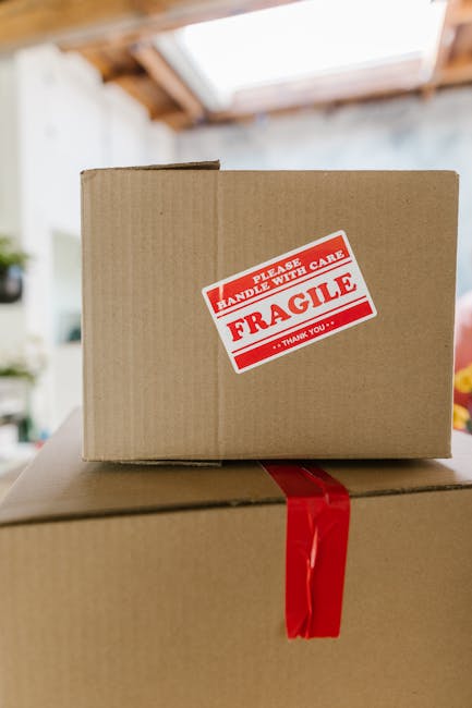 A person wearing a dark blue shirt is placing a clear glass bottle into a rectangular cardboard box filled with white packing peanuts for cushioning, preparing for a home relocation or furniture trans