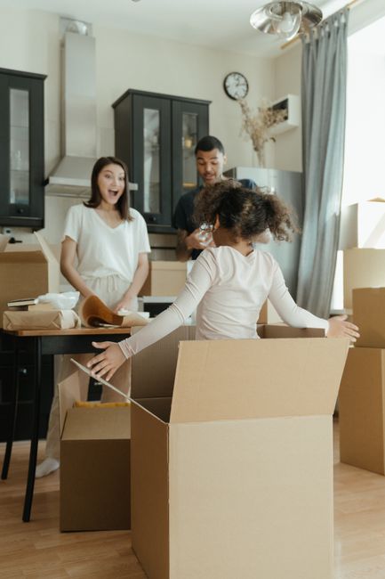 A man kneeling on the wooden floor of a room surrounded by stacked cardboard moving boxes, with some open and revealing packing materials inside. A woman and a young child are also present; the woman