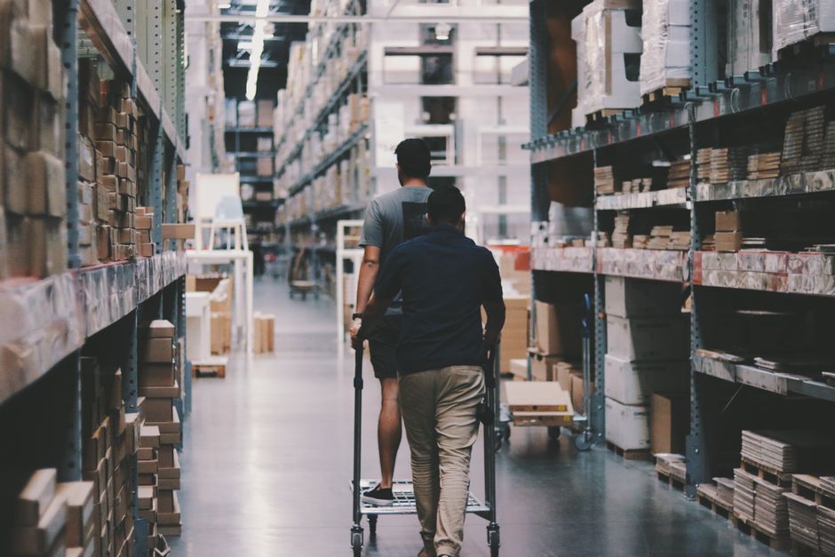A clear view inside a large warehouse or storage facility with tall metal shelving units aligned in parallel rows, filled with various boxes, containers, and packaging materials such as cardboard boxe